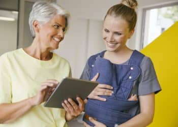 Young mother with baby carrier talking to mature woman with digital tablet in office hallway