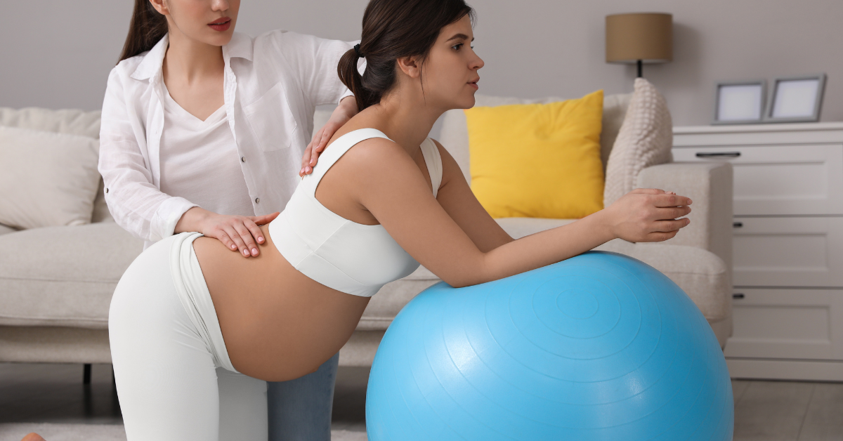 Woman in labor bent over with elbows on a birthing ball while a doula or midwife checks her back
