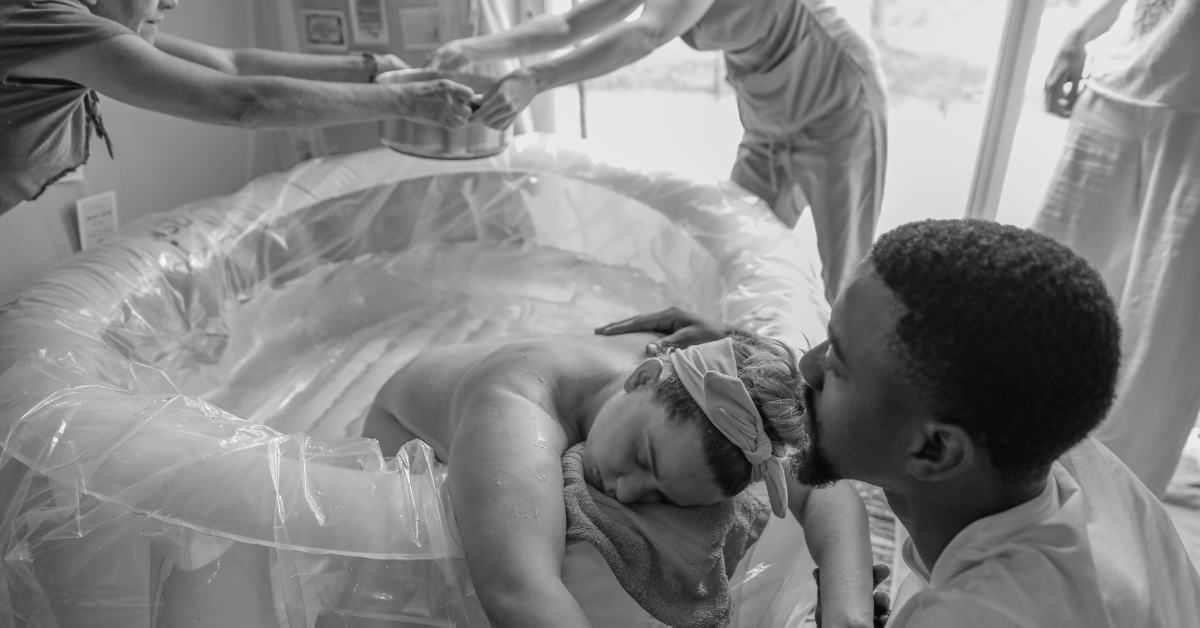 Man kissing his wife's head while she labors in a birthing pool while a midwife and a doula pass an empty bowl to be filled with more warm water for the pool