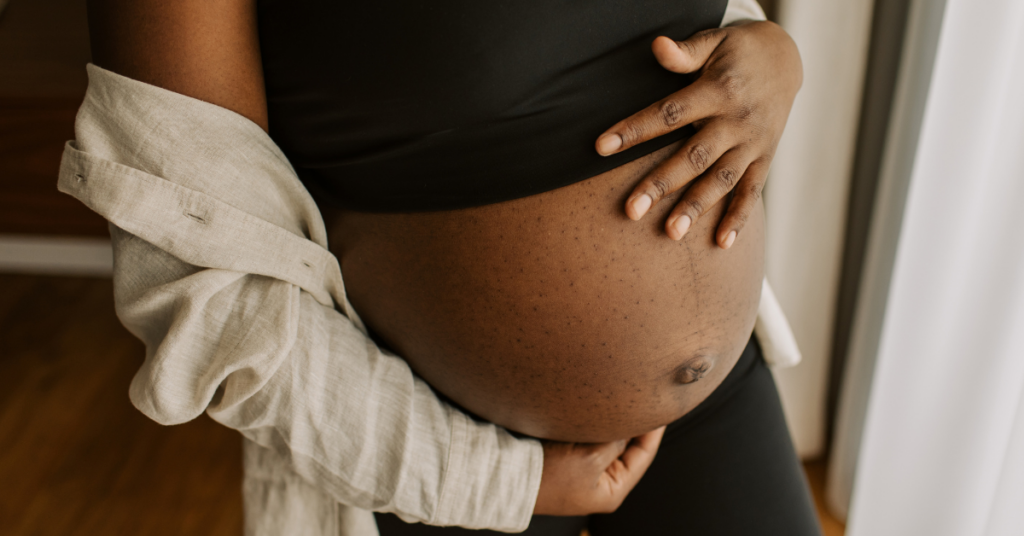 Mom at six months pregnant holding her belly in a black crop top and a sweater hanging off her arm