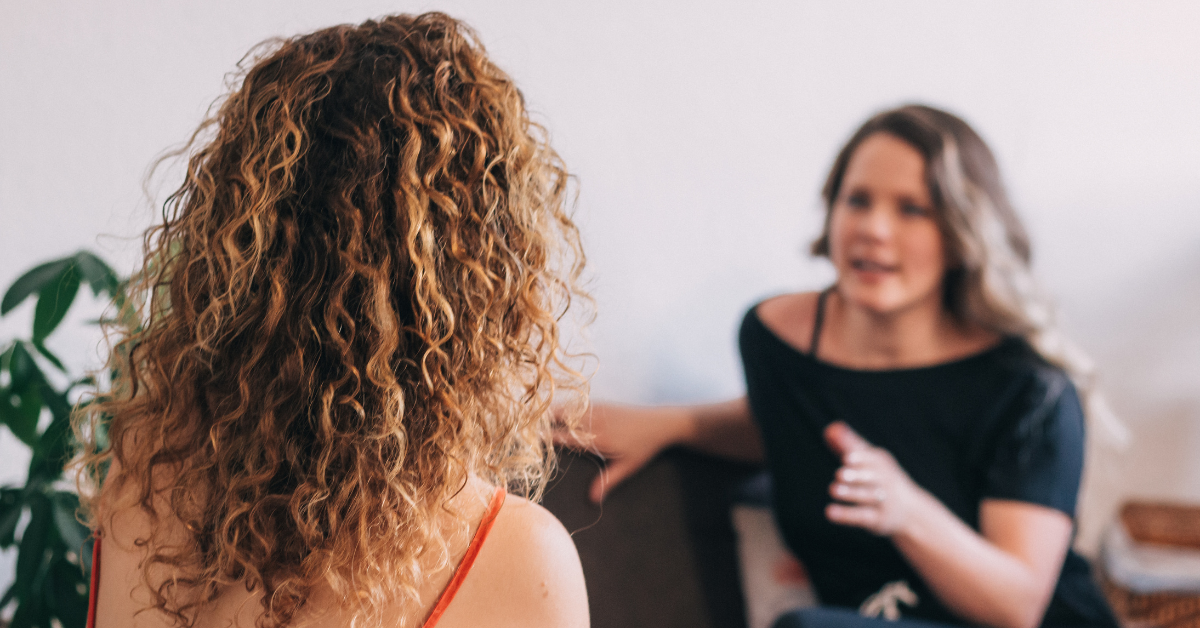 Two women in conversation: the one with curly hair is seen from behind while the other speaks to her in a casual setting.